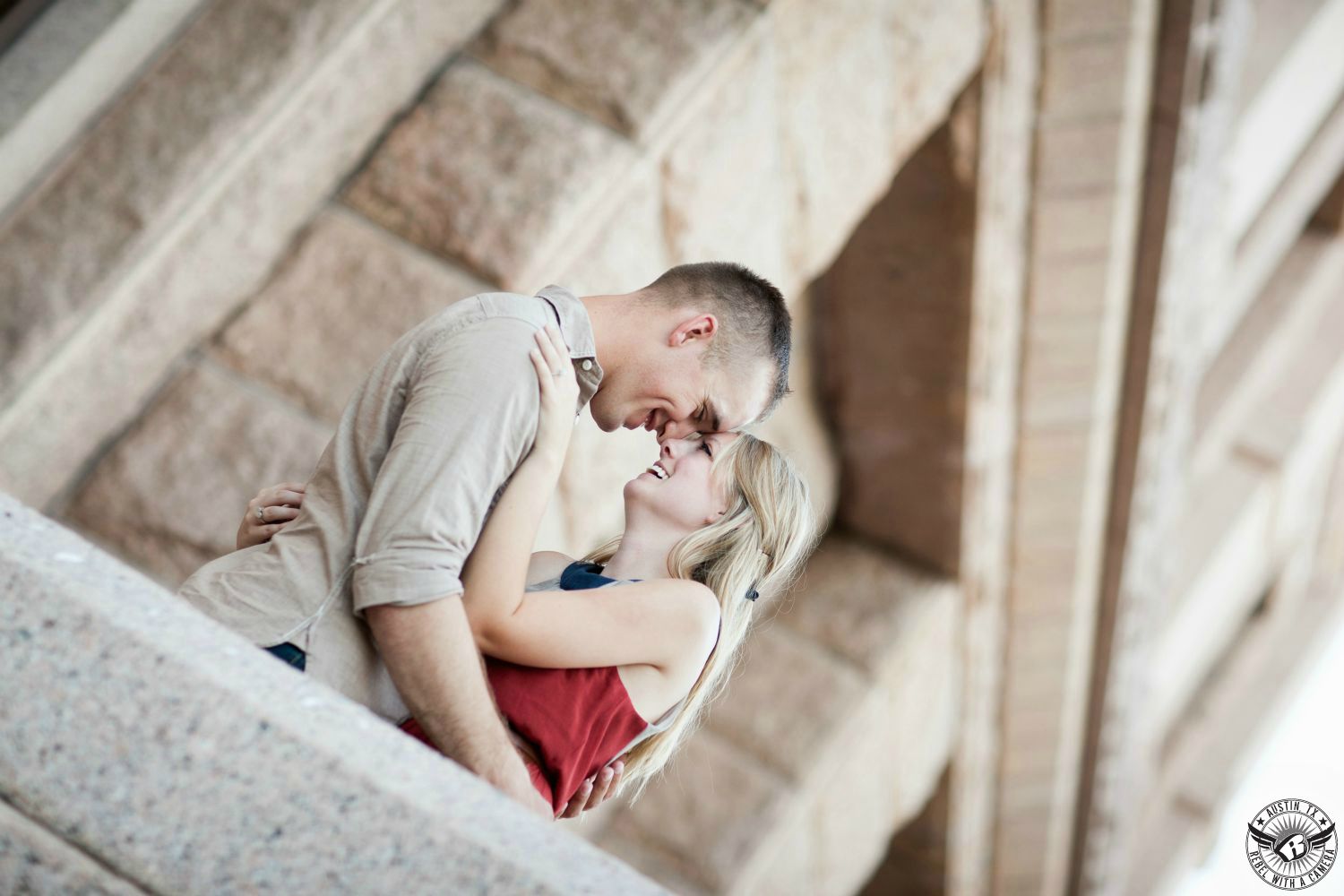 Happy blond girl in a maroon tank top looks into the eyes  of a dark haired happy guy with a tan dress shirt in front of ping granite  rough hewn stonework in this romantic engagement picture at the Texas State Capital in Austin.